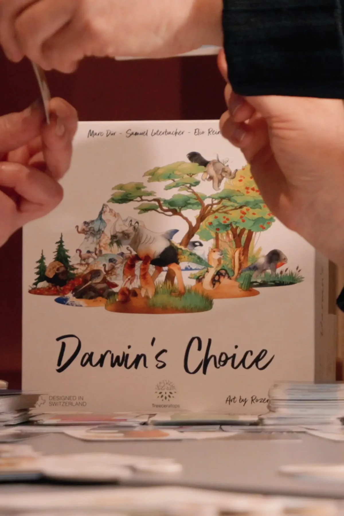 Im Hintergrund steht die Schachtel des Spieles “Darwin´s Choice”. Davor sind die Hände einiger Teilnehmender zu sehen.