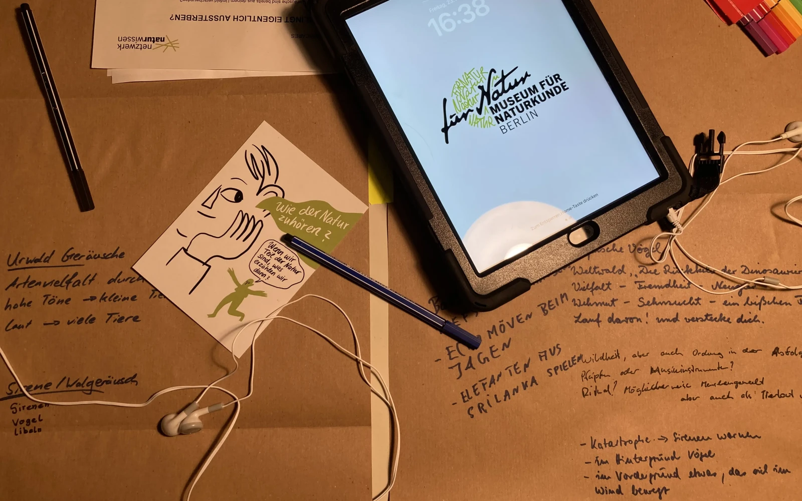 Abbildung des Arbeitsprozesses. Zu sehen sind Mitschriften, Postkarten, Papiere, Stifte, Kopfhörer sowie ein Tablet.