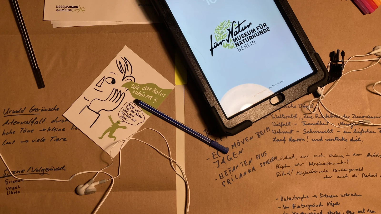 Abbildung des Arbeitsprozesses. Zu sehen sind Mitschriften, Postkarten, Papiere, Stifte, Kopfhörer sowie ein Tablet.